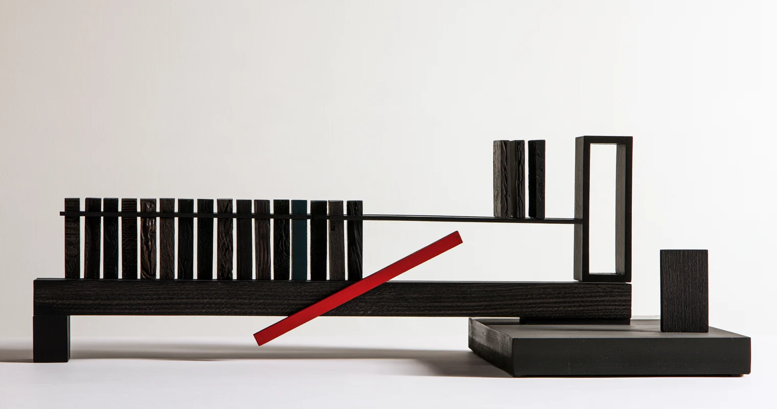 Svart trämodell där en lång bänk bär en tät rad upprättstående brickor med olika ådring, en av dem mörkt blågrön; tre brickor står avskilda intill en öppen rektangulär ram, och längst till höger står en slät bricka ensam på en låg platta medan en röd stav ligger snett över bänken.